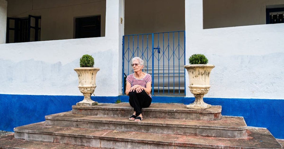 Mulher idosa sentada em degraus de concreto com corrimão azul e vasos rústicos.